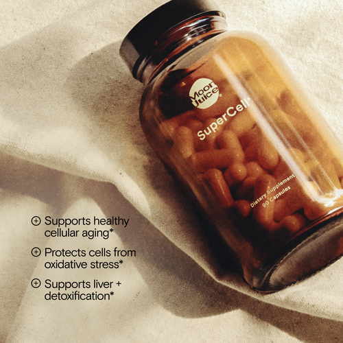 An amber glass bottle of Moon Juice SuperCell dietary supplement capsules, placed diagonally on a textured beige fabric background with soft shadows. White text on the left lists product benefits.